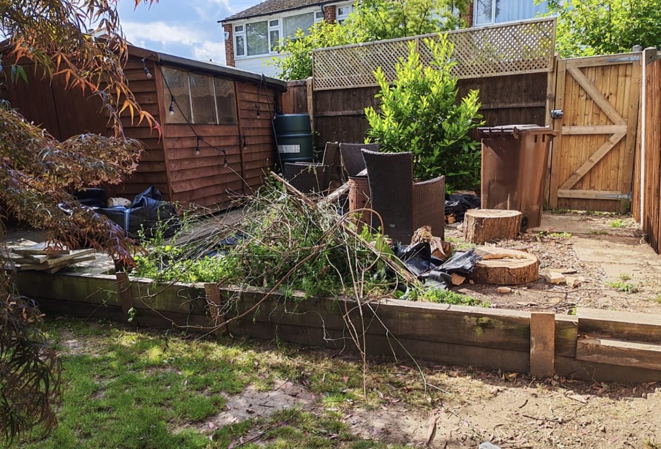 A back garden corner filled with cut branches, logs, stacked furniture and overgrown beds